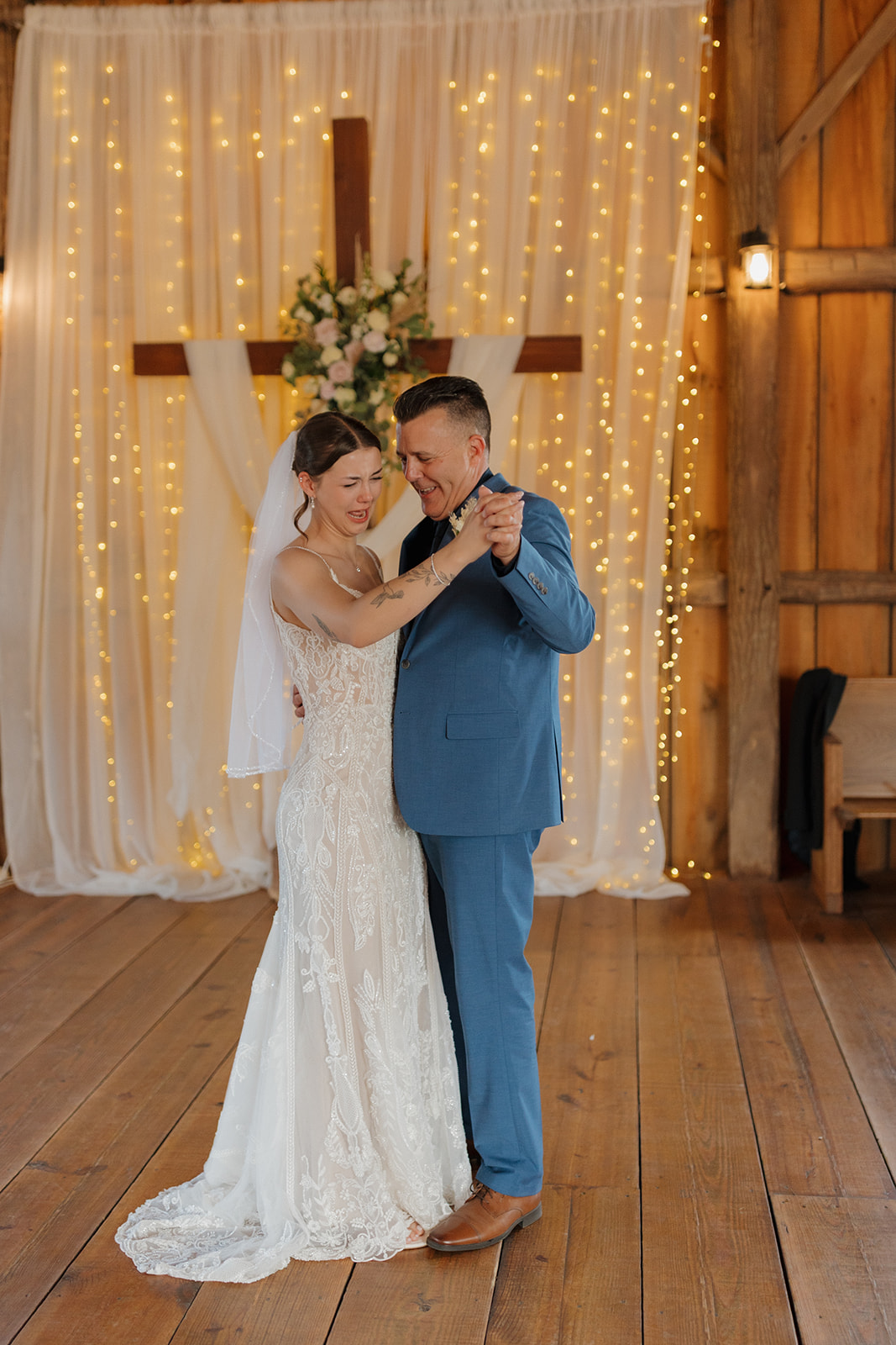 Bride dances closely with a loved one during an emotional first dance moment in a wooden barn wedding venue with twinkle lights behind them.
