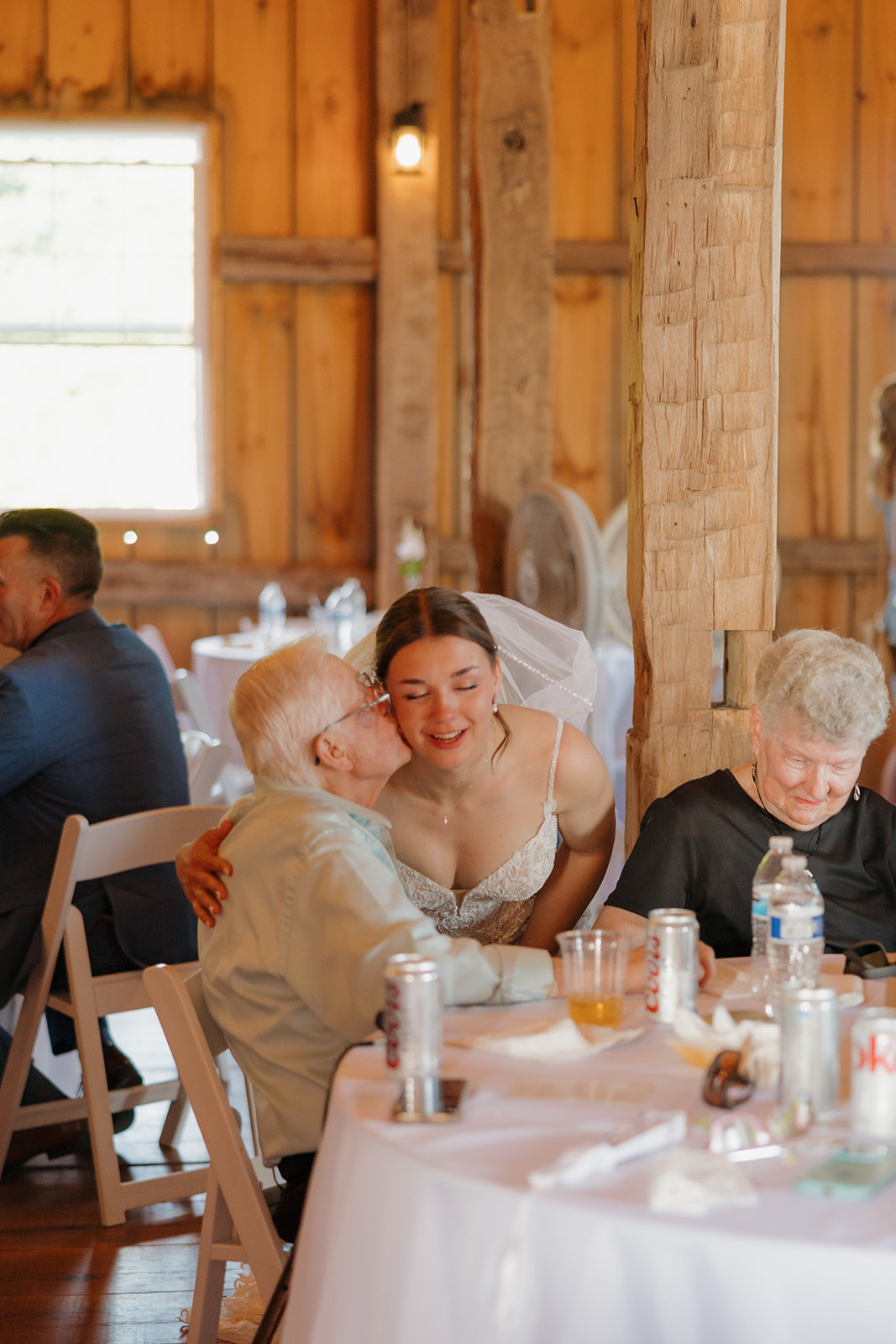 The bride leans in to hug an elderly family member at a reception table, sharing a quiet, emotional moment during a Wisconsin barn wedding.