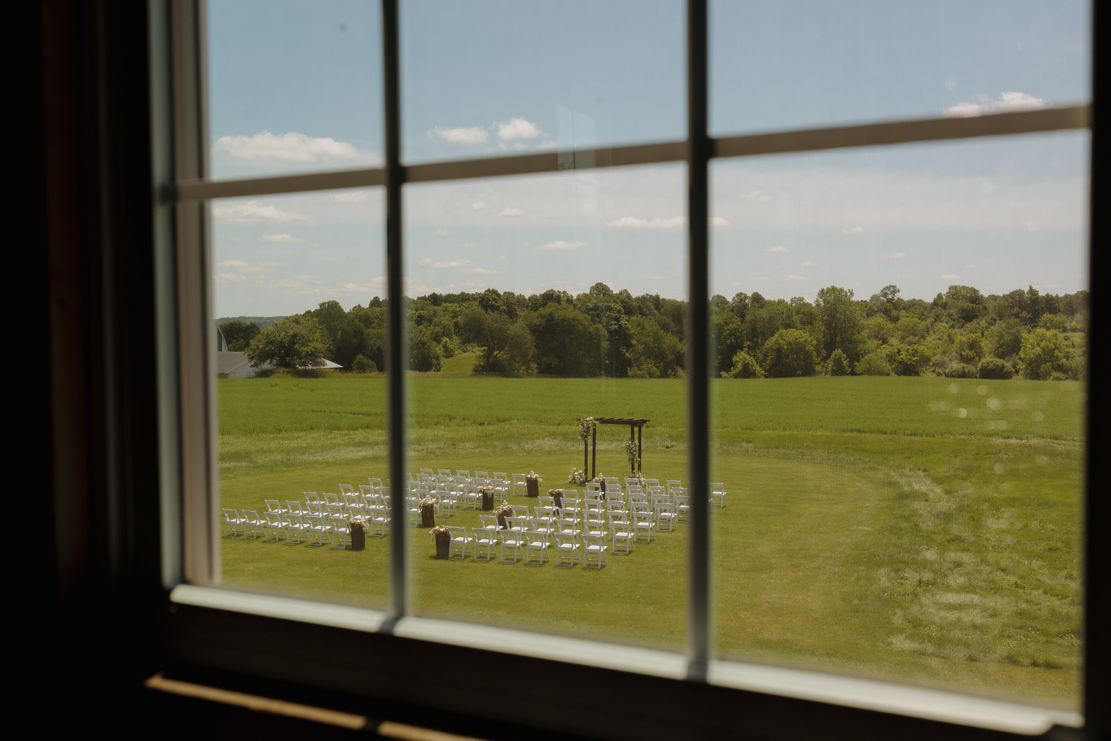 Outdoor wedding ceremony setup viewed through a window overlooking green fields, captured by a madison wisconsin wedding photographer.