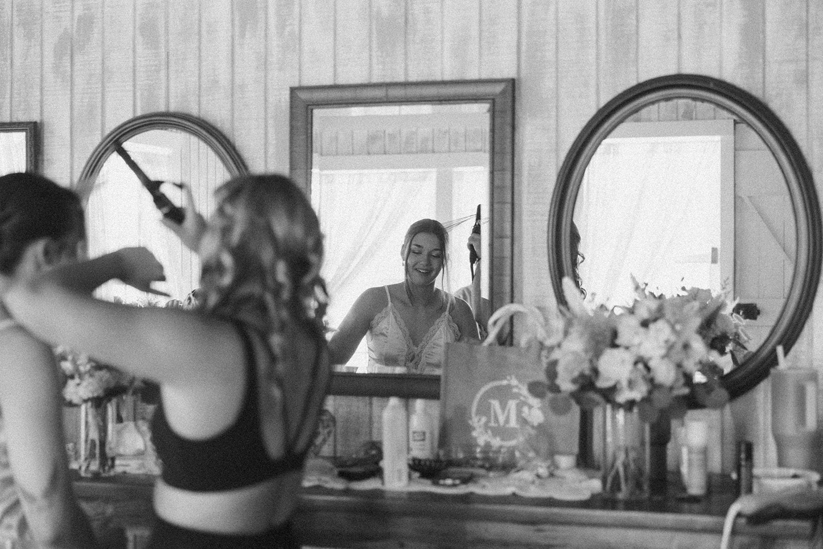 Bride smiling in the mirror while bridesmaids curl her hair during wedding morning preparations, captured by a madison wisconsin wedding photographer.