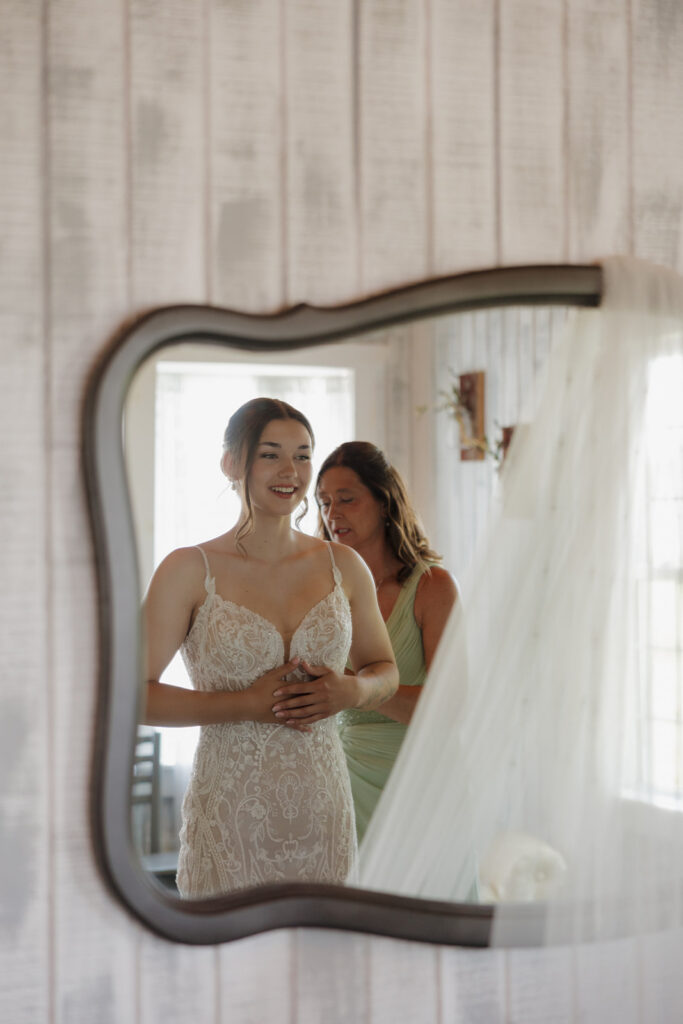 Bride smiling at her reflection while getting ready in a lace wedding dress, photographed by a madison wisconsin wedding photographer.