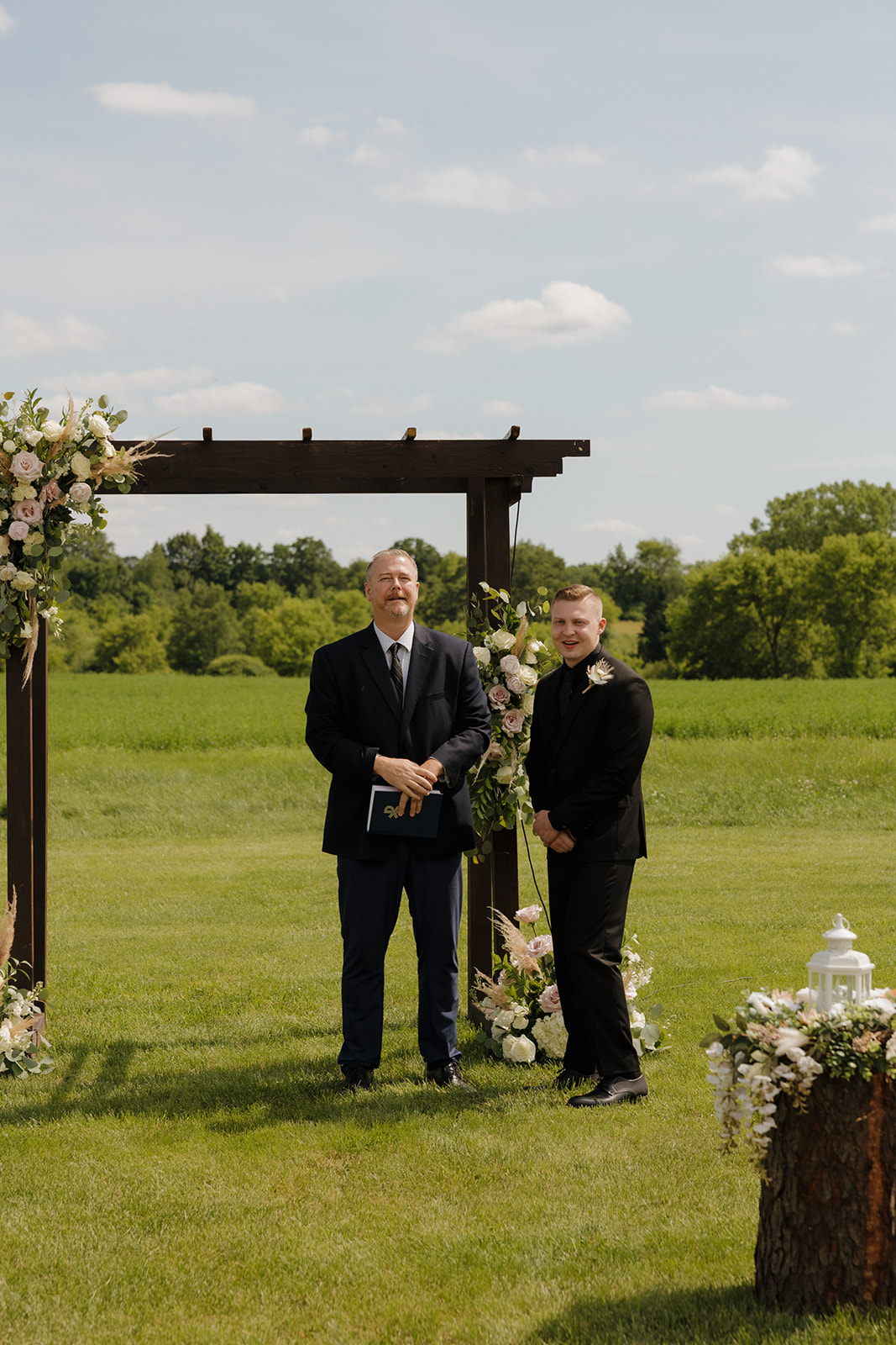 Outdoor wedding ceremony setup with groom and officiant under a floral arch in a green field, photographed by a madison wisconsin wedding photographer.