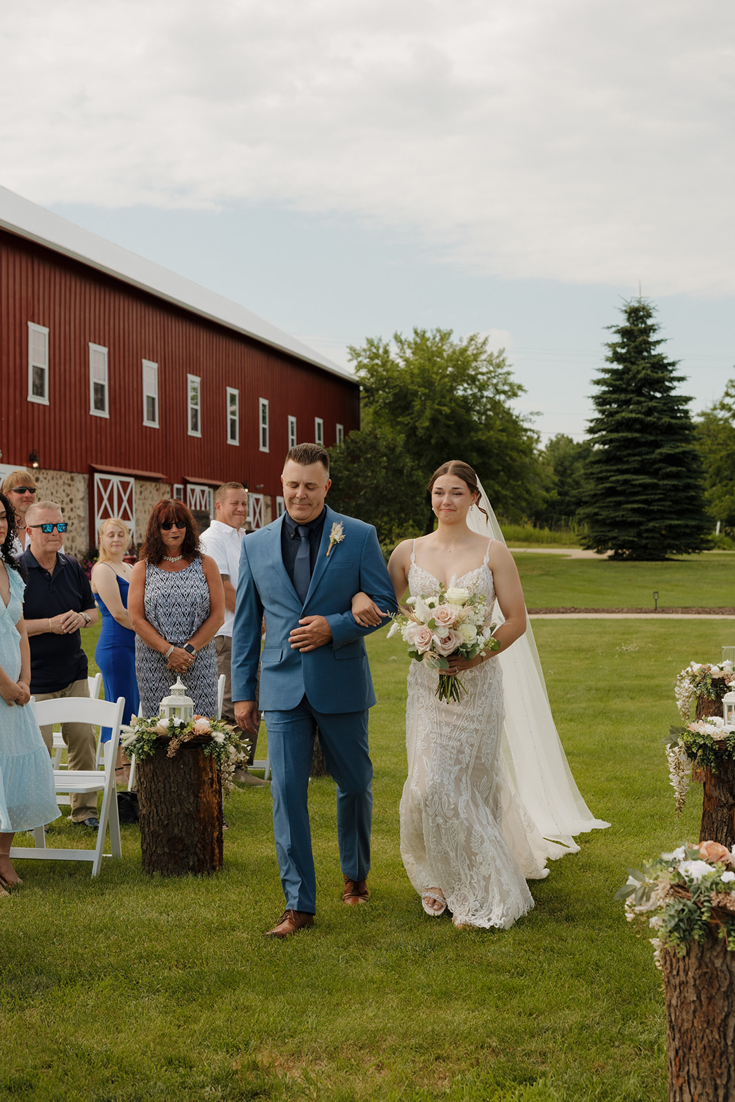 Bride walking down the aisle with her father in front of a red barn at an outdoor wedding, captured by a madison wisconsin wedding photographer.