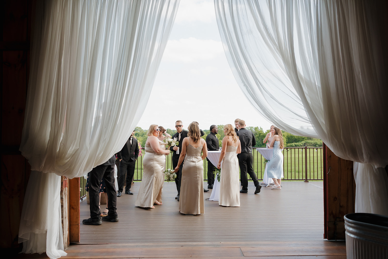 Wedding guests mingle and chat during cocktail hour on a wooden deck, framed by flowing white drapery and open countryside views, captured by a madison wisconsin wedding photographer.
