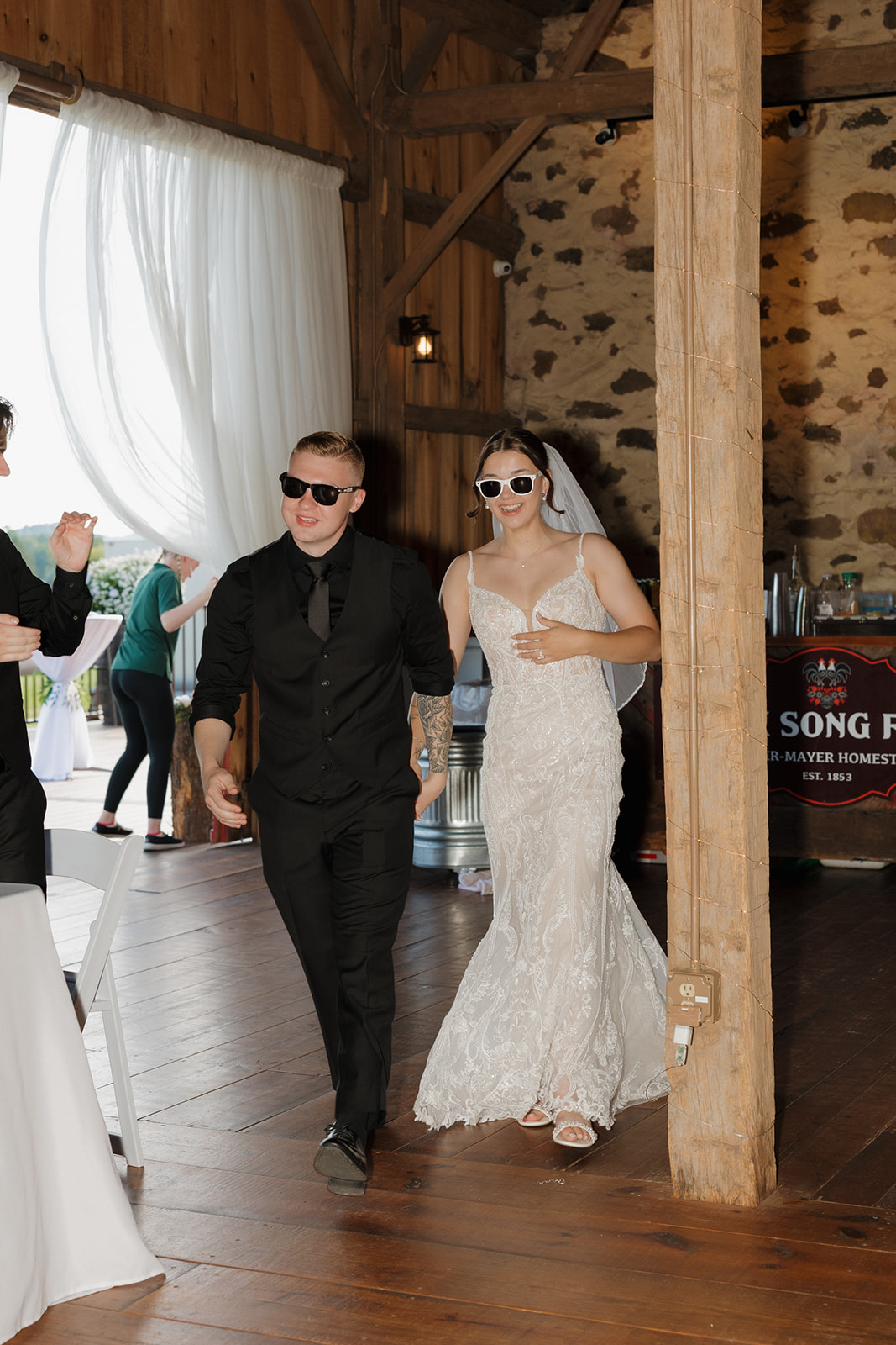 Bride and groom wearing sunglasses make a playful reception entrance into a rustic barn, hand in hand and smiling as guests cheer.