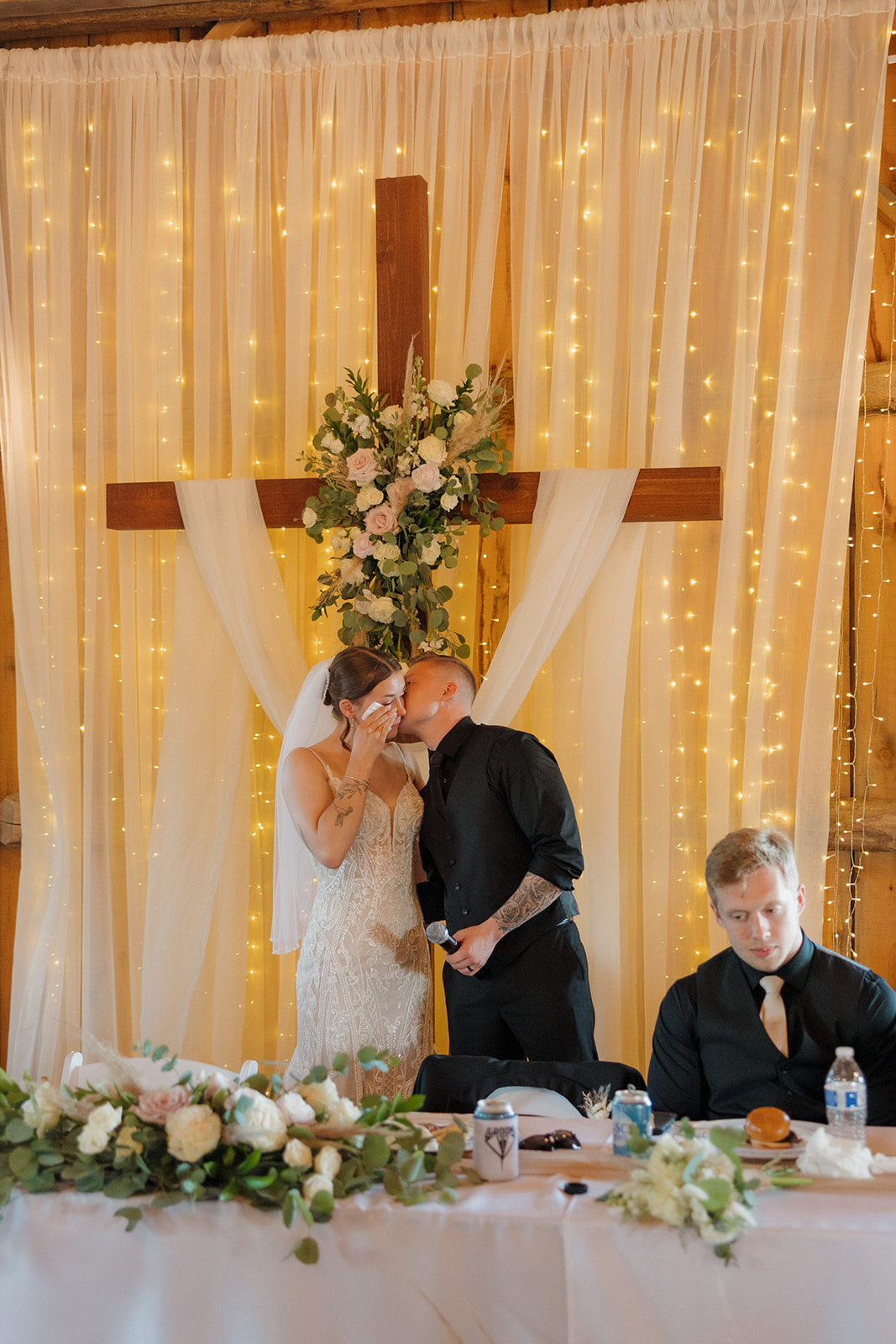 The groom kisses the bride’s cheek at the head table as she laughs, surrounded by florals and warm string lights, photographed by a madison wisconsin wedding photographer.