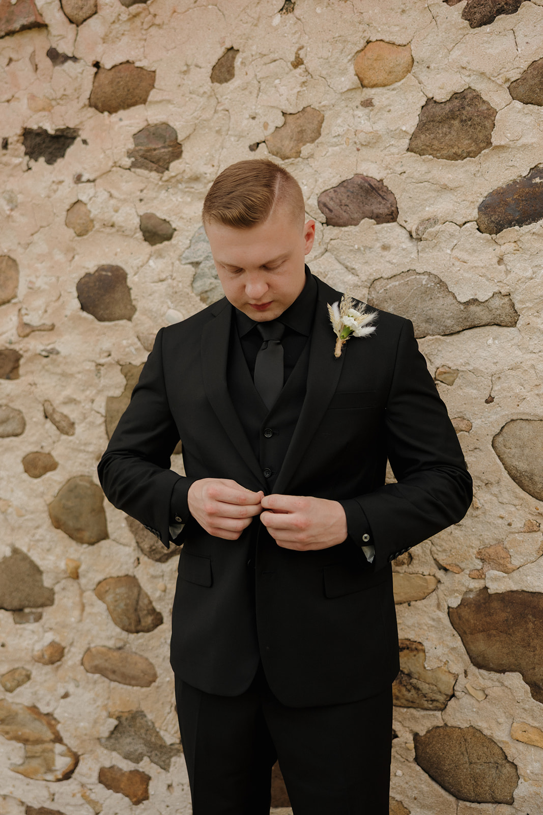 Groom buttoning his black suit jacket in front of a textured stone wall, photographed by a madison wisconsin wedding photographer.