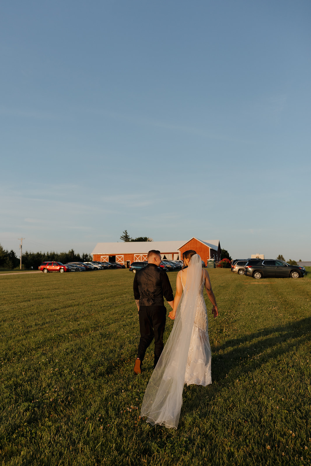Bride and groom walking hand in hand across a field at golden hour near a red barn, captured by a madison wisconsin wedding photographer.