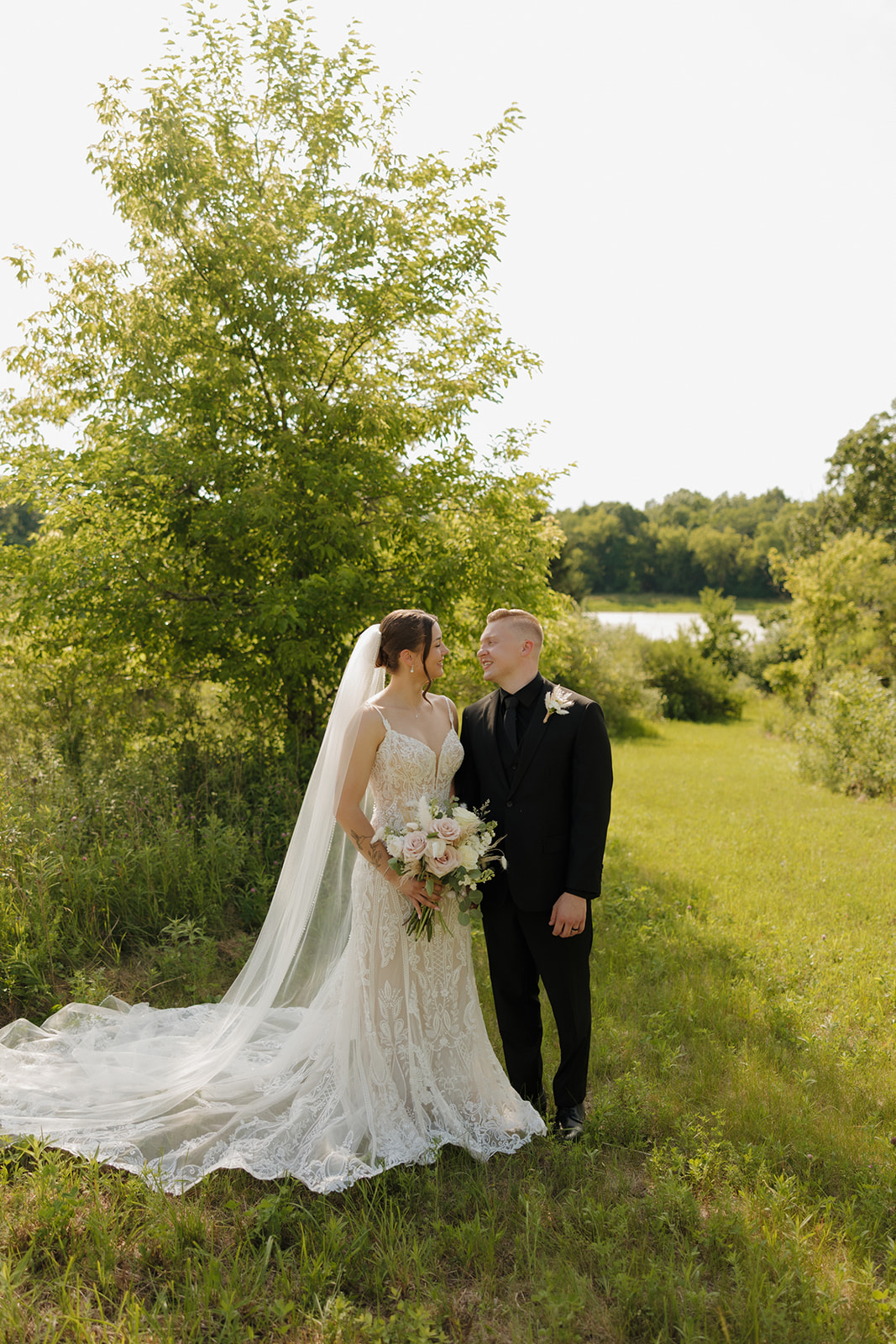 Bride and groom standing together in a sunlit field with trees and water in the background, photographed by a madison wisconsin wedding photographer.