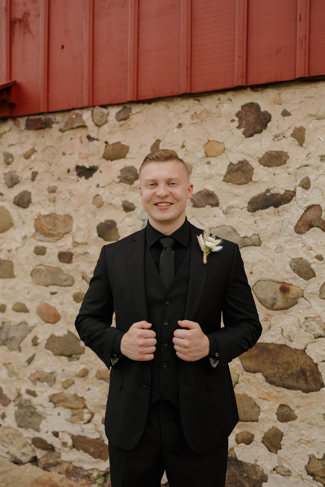Groom in a black suit smiling in front of a rustic stone barn wall, captured by a madison wisconsin wedding photographer.