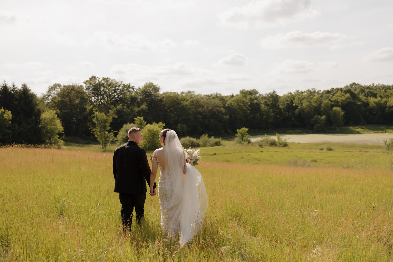 The bride and groom walk away together through tall grass and open fields, seen from behind during a peaceful outdoor wedding portrait at golden hour.
