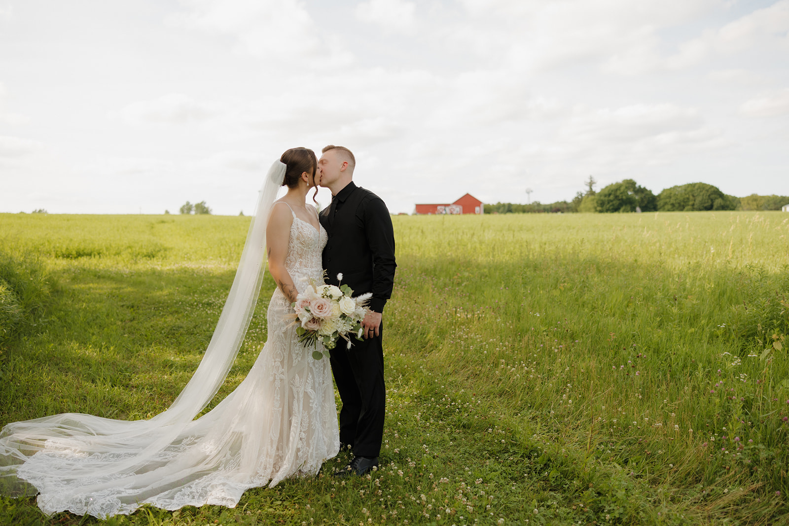 Bride and groom sharing a kiss in a green field with a red barn in the distance, photographed by a madison wisconsin wedding photographer.