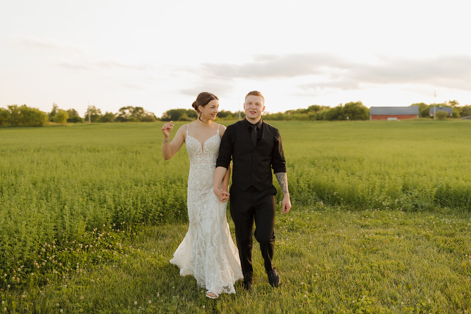 Bride and groom walk hand in hand through a green field at sunset, smiling at each other during golden hour portraits with a madison wisconsin wedding photographer.