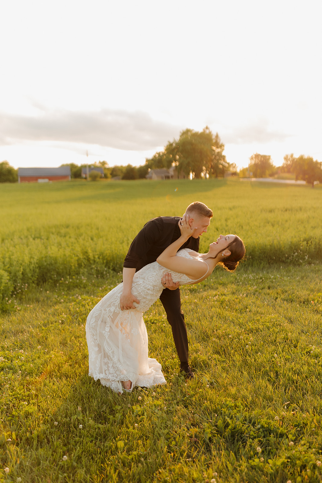 The groom dips the bride into a kiss at golden hour in an open field, surrounded by soft sunlight and summer greenery.