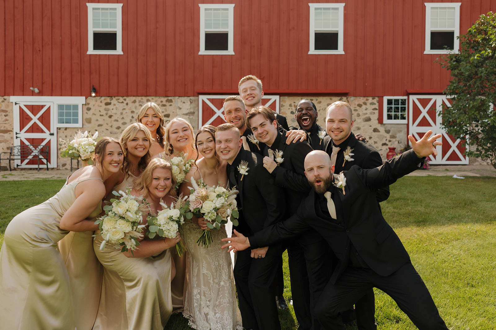 The full wedding party gathers closely on the lawn in front of a red barn, smiling and celebrating together during a joyful group portrait.