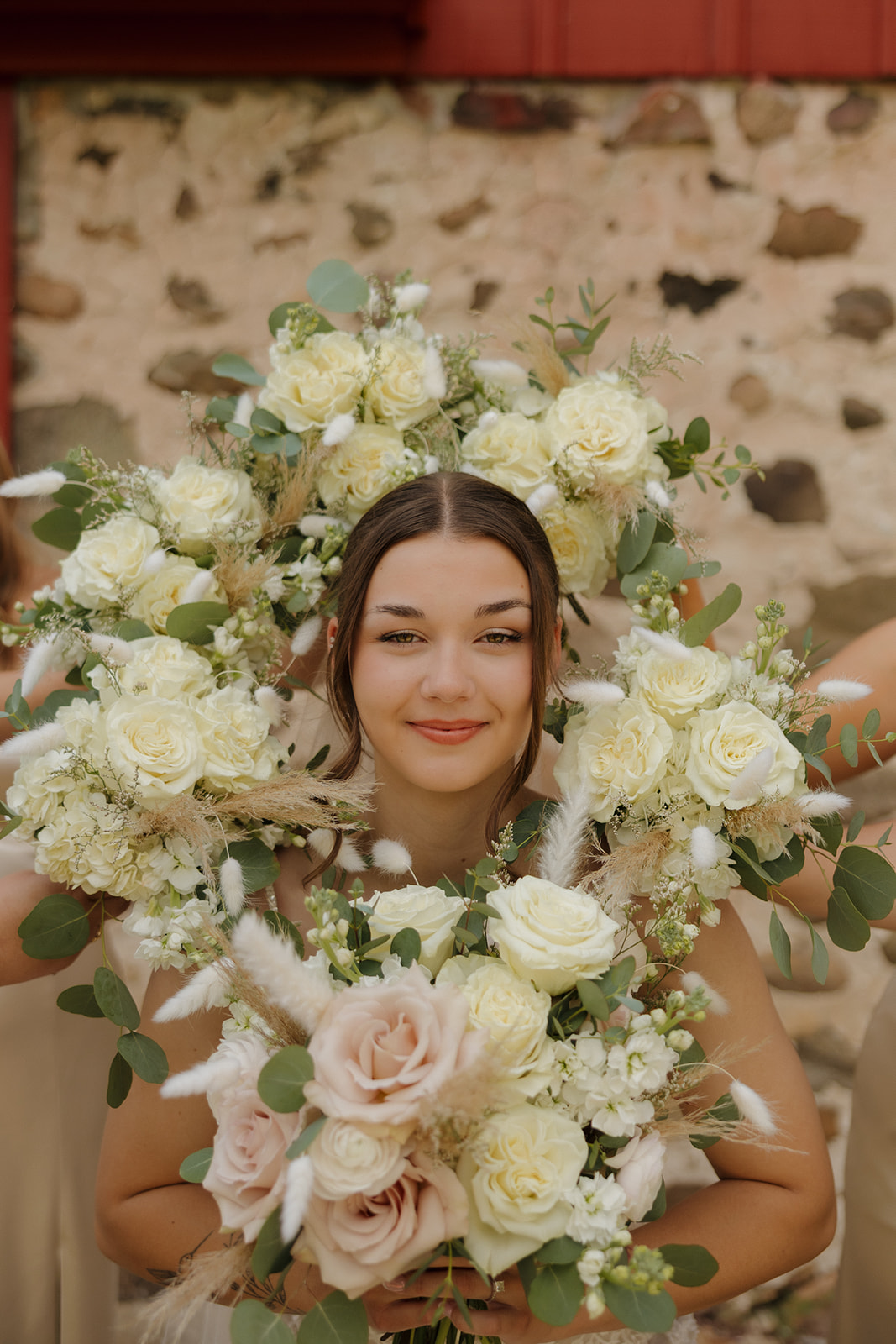 A bridesmaid smiles while holding layered floral bouquets around her face, creating a playful portrait moment outside a rustic stone-and-barn backdrop, photographed by a madison wisconsin wedding photographer.
