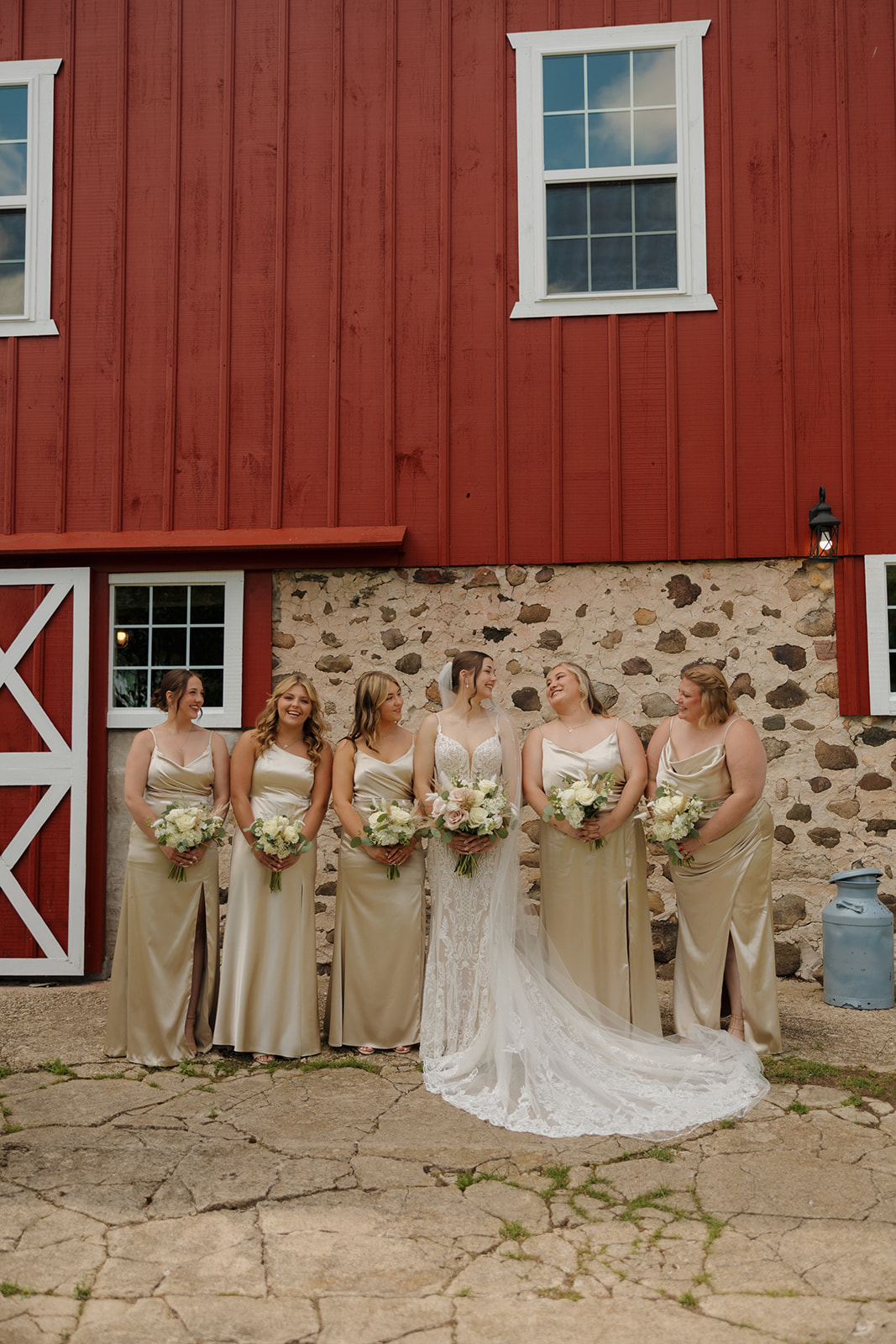 Bride stands laughing with her bridesmaids in champagne dresses outside a red barn, holding bouquets and sharing a relaxed pre-ceremony moment, photographed by a madison wisconsin wedding photographer.