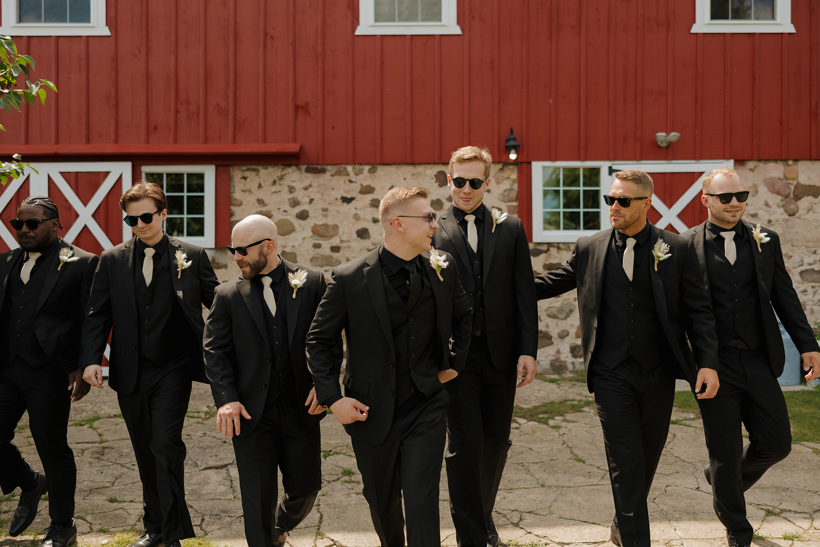 The groom and groomsmen walk together in black suits and sunglasses outside a red barn, laughing and talking during wedding party portraits captured by a madison wisconsin wedding photographer.