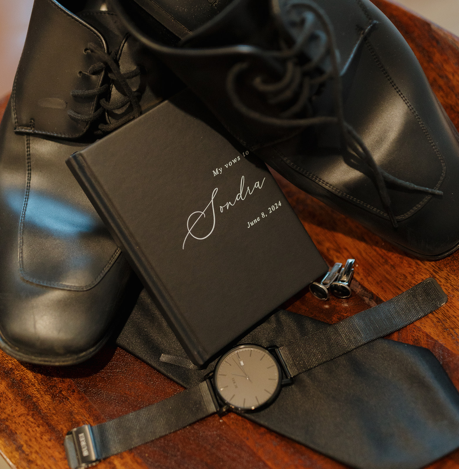 Groom details with black shoes, watch, and vow book on wooden table
