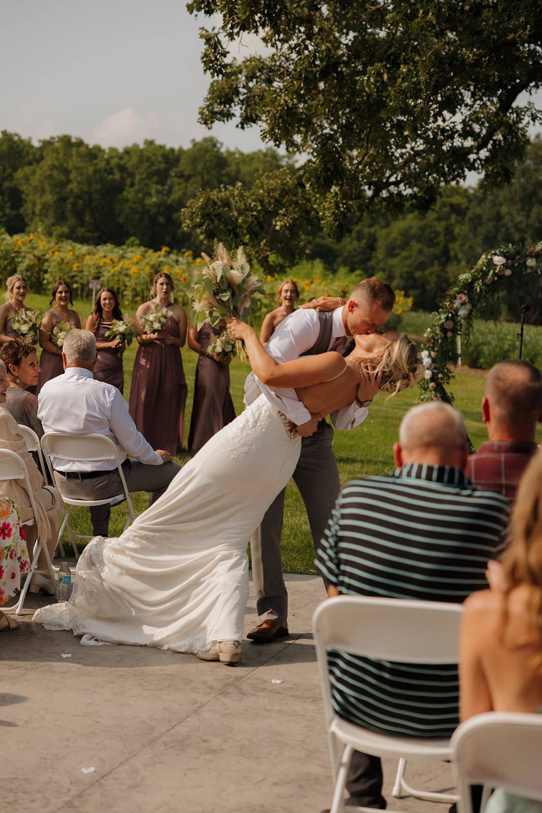 Groom dips the bride dramatically for a kiss while guests cheer them on outdoors.