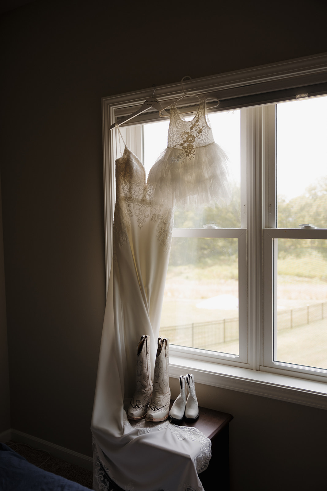 Bride and toddler’s dresses hanging by the window with boots—an intentional detail shot for a wedding photography checklist.