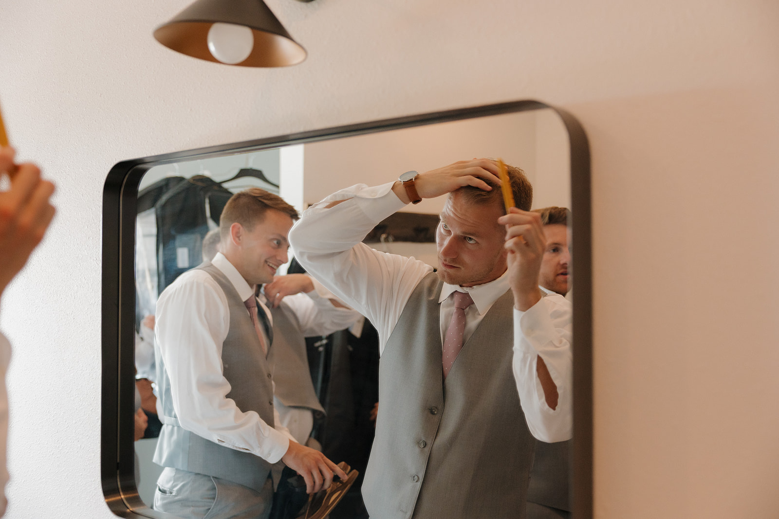Groom adjusting his hair in the mirror while groomsmen get ready in the background.