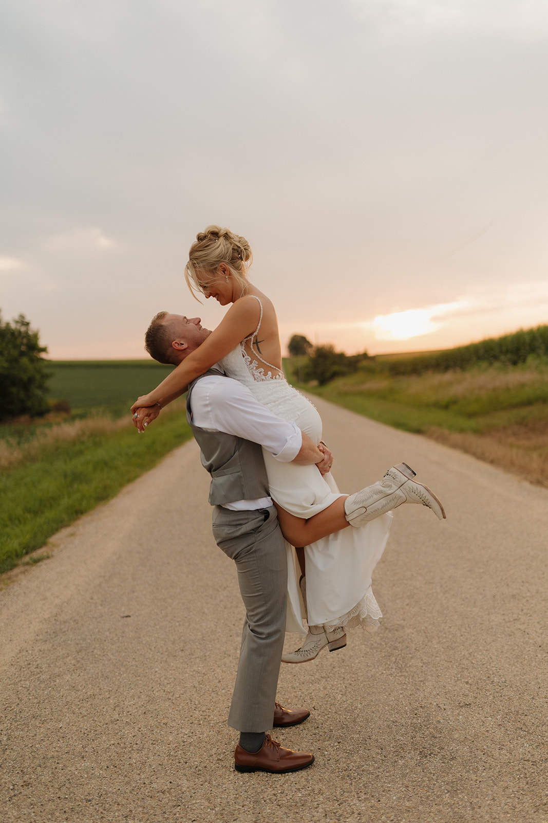 Groom lifts the bride in celebration on a rural road at sunset, her boots adding a fun western twist.