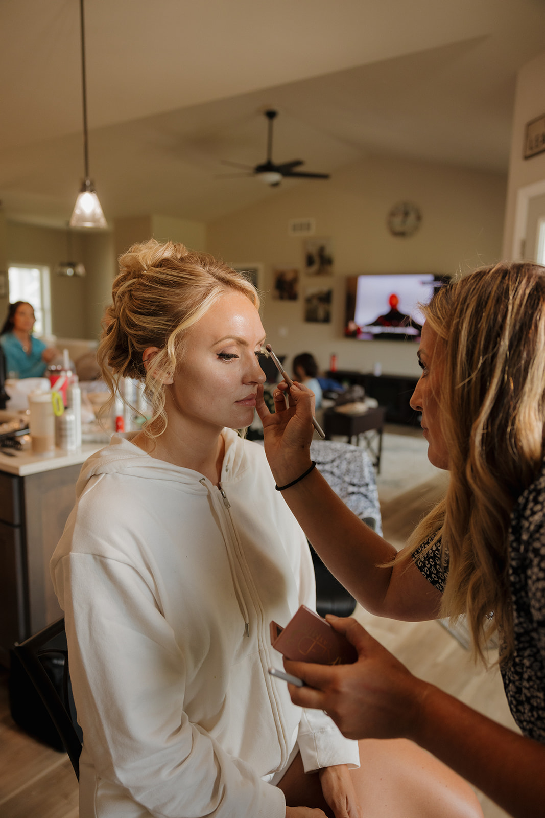 Bride getting makeup done in a cozy kitchen surrounded by her people.