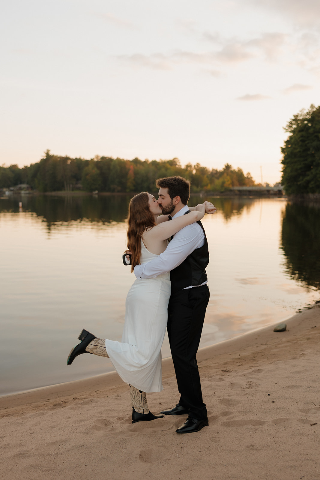 Couple kissing on a quiet lakeshore beach at sunset after their intimate wedding celebration.