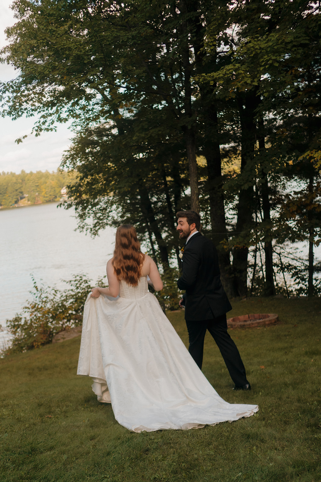 Bride and groom walking together along a lakeside lawn surrounded by tall trees during romantic wedding portraits.