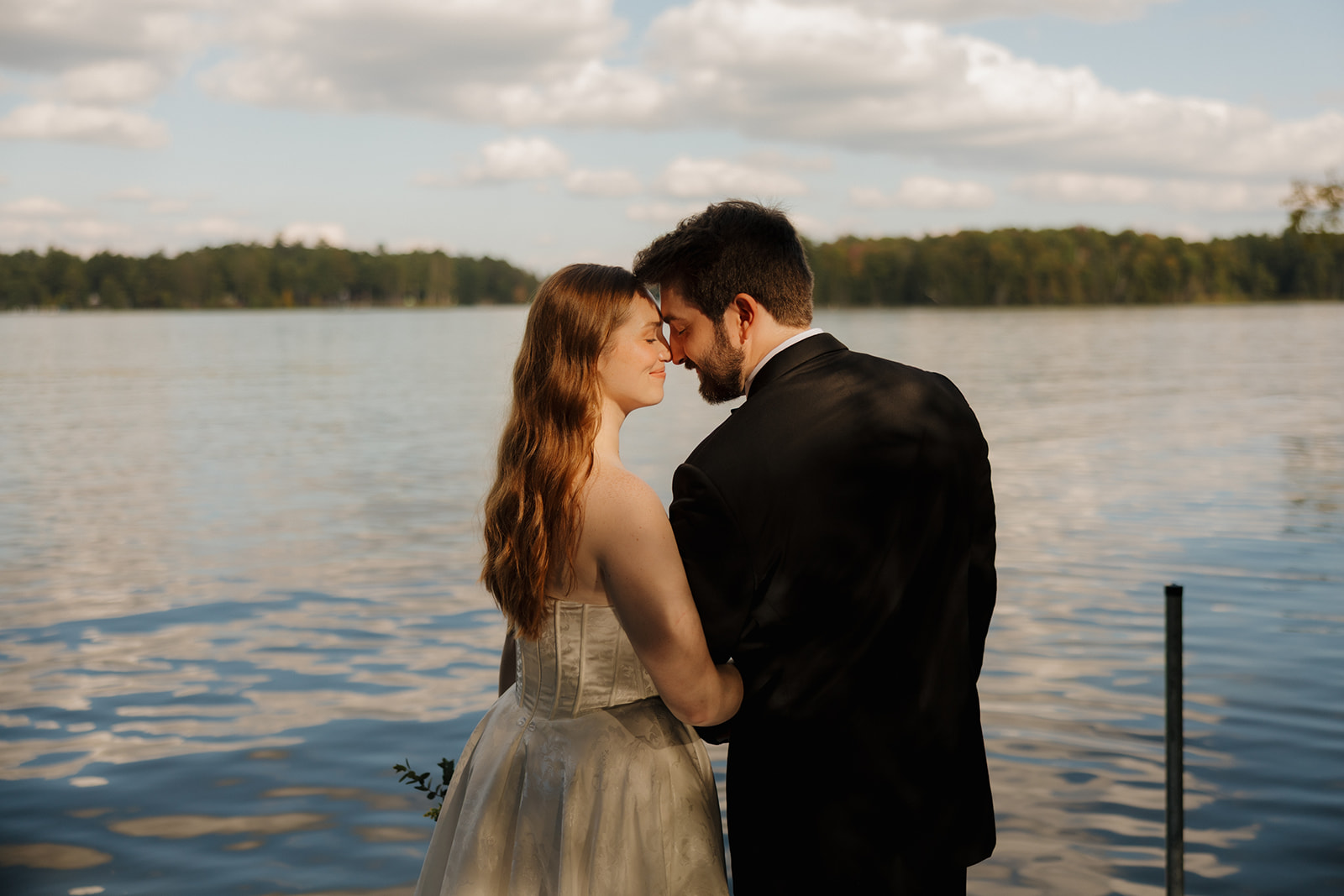 Bride and groom standing together along the water at sunset during an outdoor wedding portrait