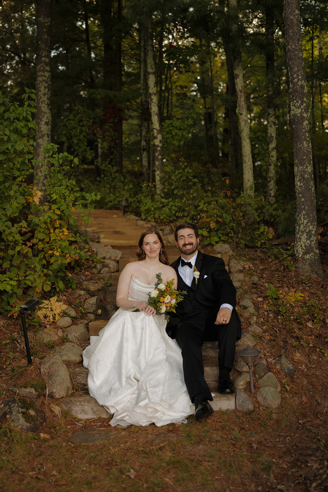 Bride and groom sitting together on stone steps surrounded by forest at an outdoor wedding venue.
