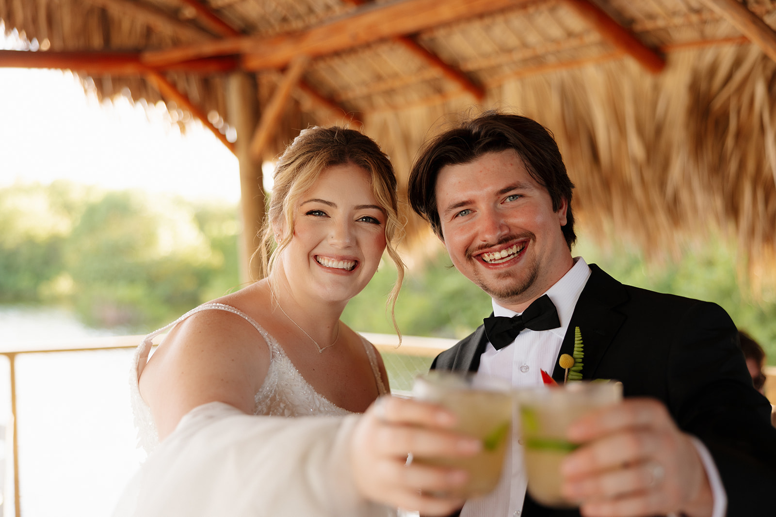 Bride and groom smiling and toasting cocktails together during a tropical wedding in Florida under a thatched pavilion near the water.
